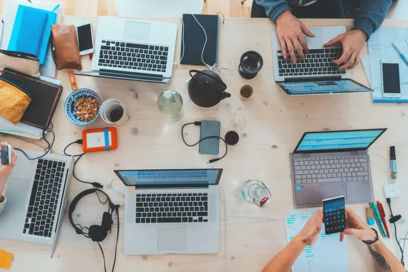 Overhead view of a wooden desk with multiple laptops, phones, headphones, and snacks, suggesting a collaborative workspace.