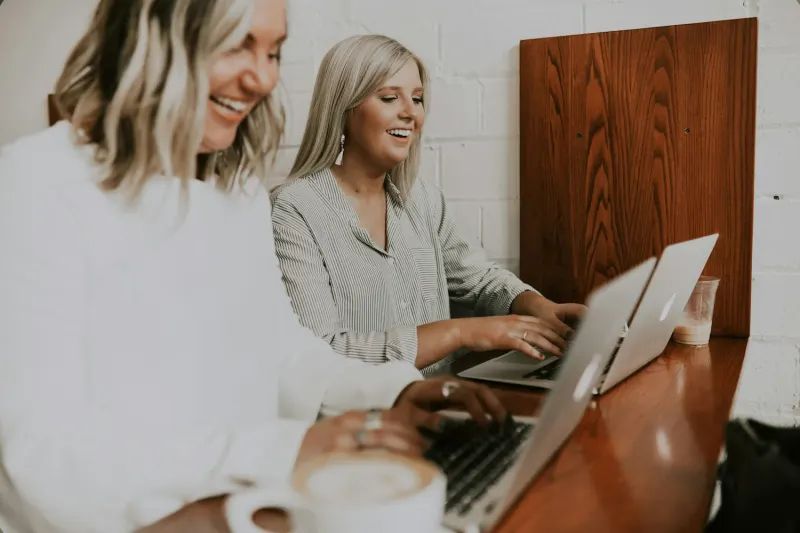 Two women smiling and working on laptops at a coffee shop with a latte in the foreground.