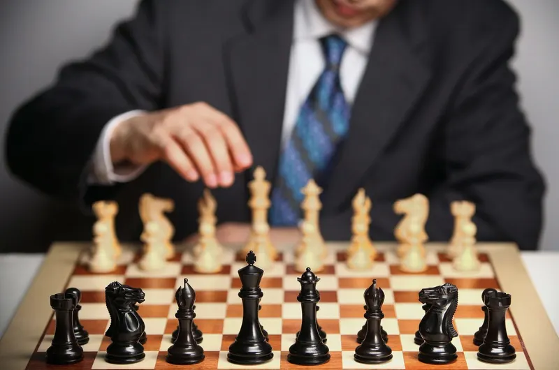 A person in a suit and tie is about to move a white chess piece across the board, black pieces in the foreground.