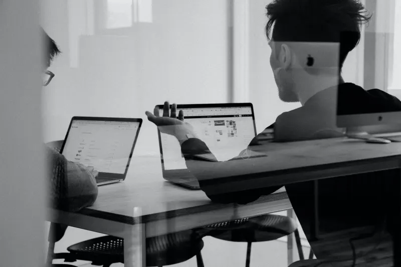 Two people sitting at a table with laptops, engaged in a discussion.