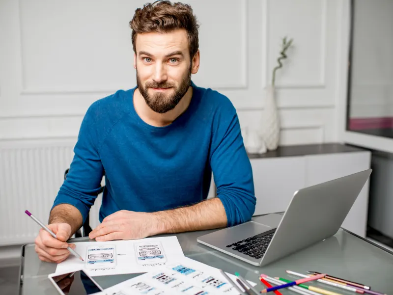 Man in a blue sweater at a desk holding a pencil and looking at wireframe mockups for a mobile app, a laptop on the desk.