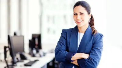 Smiling businesswoman in a dark blue suit jacket with arms crossed, standing in an office with computers in the background.