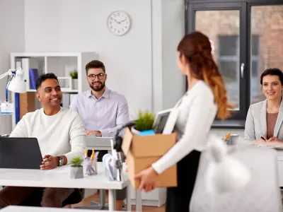 A woman carries a box of office supplies as a new employee starts a job while colleagues smile.