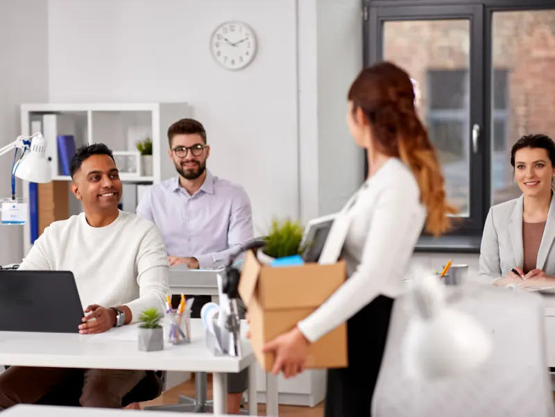 A woman carries a box of office supplies as a new employee starts a job while colleagues smile.