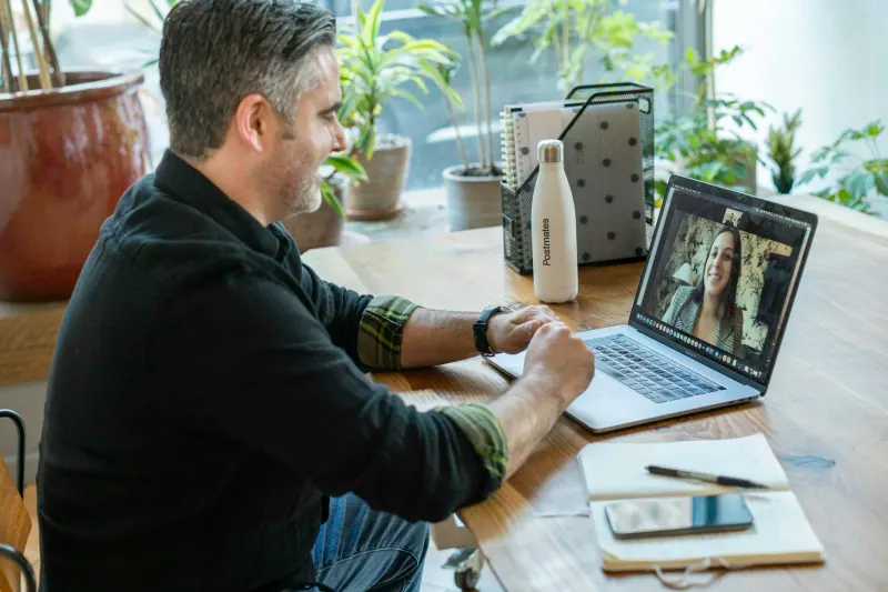 A man smiles while on a video call with a coworker on his laptop at a wooden desk — the kind of interview conversation where SDR candidates are judged on speed, follow-up and channel fluency.