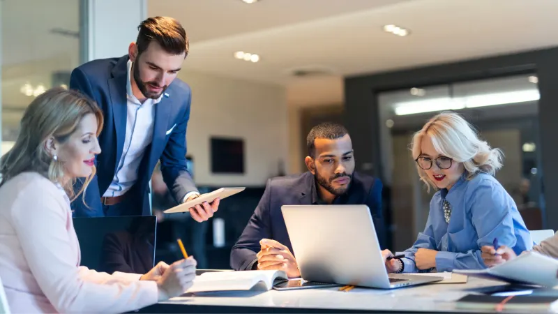 Four professionals in a meeting, two standing and two seated, looking at laptops and documents at a table.