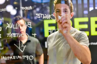 A young man in a gray t-shirt writes on a glass board covered in white text and symbols, another man blurred in the background.