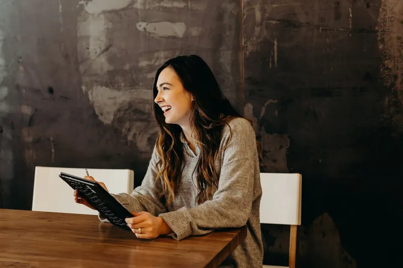 A woman with long wavy brown hair in a gray sweater laughs while holding a tablet and stylus at a wooden table.