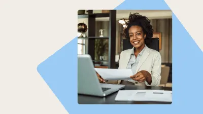 Smiling Black woman in a light blazer sits at a desk with a laptop and papers, holding a document.