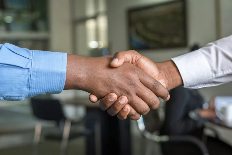 Two hands clasping in a handshake; one person wears a blue dress shirt, the other a white dress shirt.