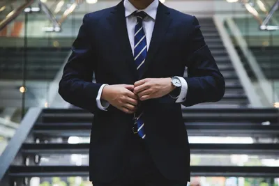 A man in a dark suit and striped tie fastens his jacket buttons while standing on stairs.