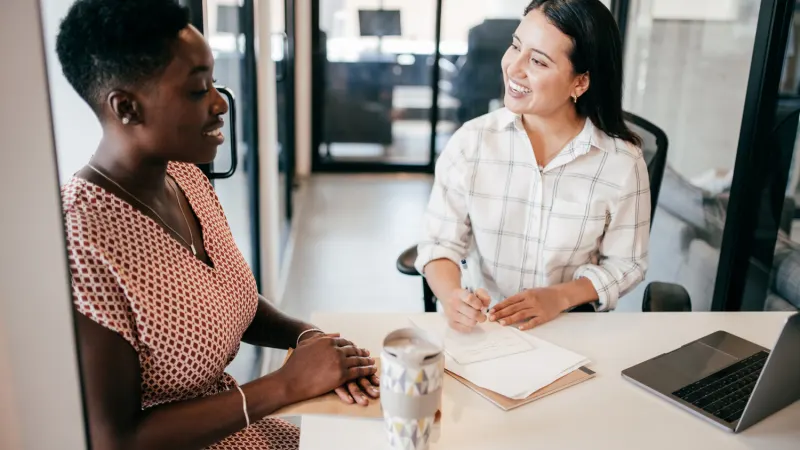 Two women during a business meeting, one taking notes as the other looks on smiling.