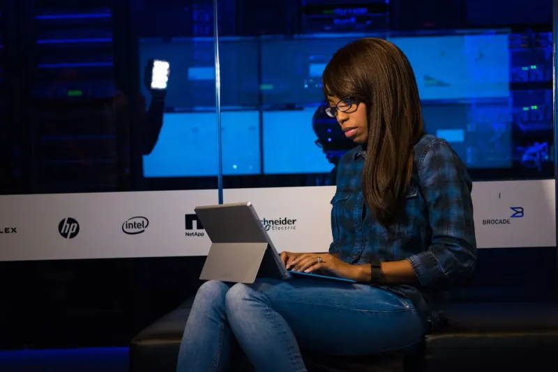 Woman in glasses and a plaid shirt types on a tablet in front of server racks and company logos.