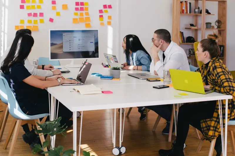 A diverse group of five people sit around a white table, focused on laptops and a large monitor displaying project management software.