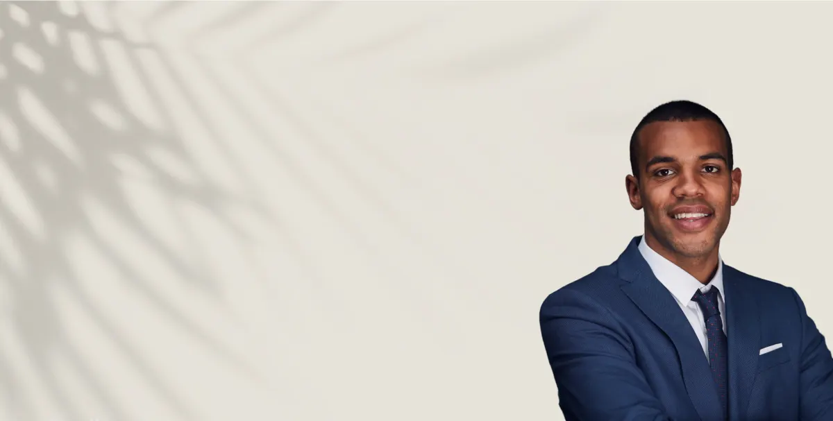 A smiling professional in a navy suit standing against a light wall patterned with palm-frond shadows