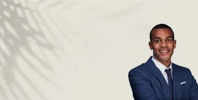 Man in a blue suit and white shirt smiling with arms crossed, against a neutral background with palm-leaf shadows.