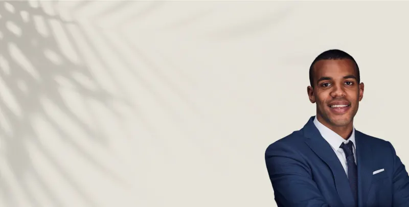 A smiling professional in a navy suit standing against a light wall patterned with palm-frond shadows