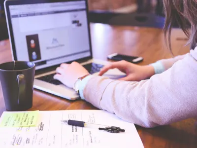 Woman wearing a beige sweater types on a laptop next to a mug and notes.