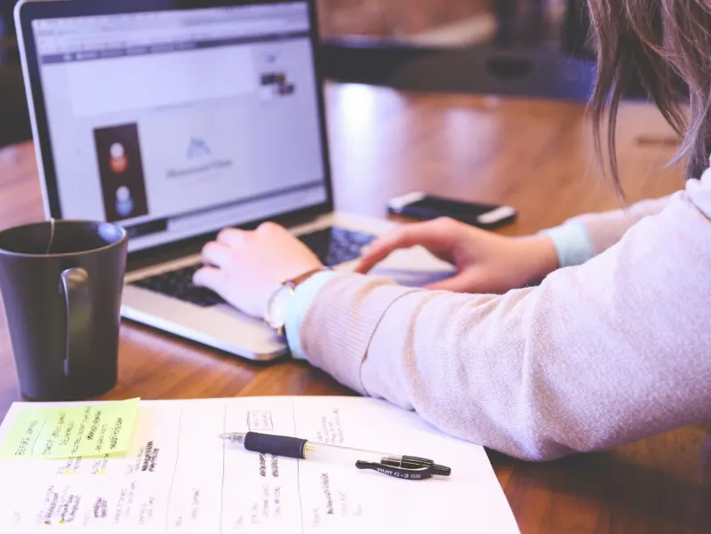 Woman wearing a beige sweater types on a laptop next to a mug and notes.