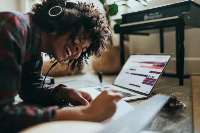 A smiling person with headphones lies on the floor writing in a notebook next to a laptop displaying audio editing software.