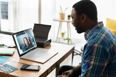 A Black man in a blue plaid shirt smiles while video conferencing with a woman on his laptop.