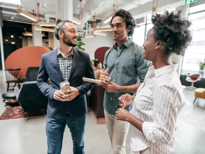 Three professionals in a modern office converse; one man holds a patterned mug, another a notebook, and a woman smiles.
