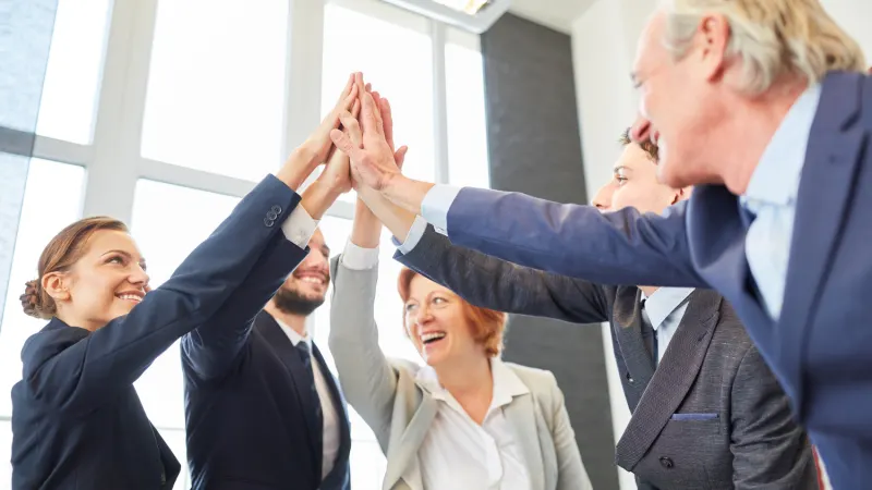 A group of diverse business people in suits high-fiving each other with smiles, celebrating.