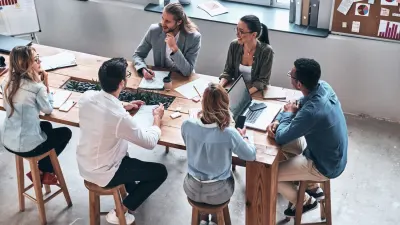 Six people sit around a modern wooden conference table, engaged in discussion with laptops and papers.