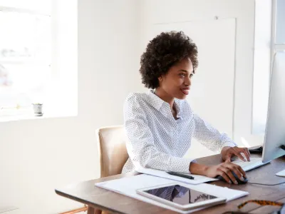 Smiling woman with afro hair in a polka dot shirt typing on a computer keyboard at a desk.