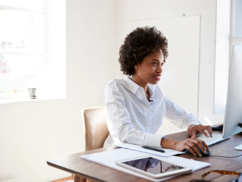 Smiling woman with afro hair in a polka dot shirt typing on a computer keyboard at a desk.