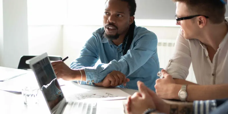 A Black man in a denim shirt speaks animatedly while pointing at a laptop screen during a meeting; a man in glasses and a white shirt sits opposite him.