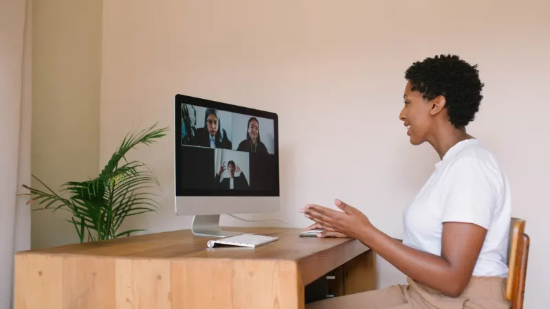 A Black woman in a white t-shirt smiles and gestures while on a video call with three colleagues on her screen.