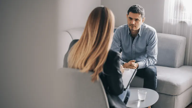 A man sits on a couch looking at a therapist who is taking notes, in a bright room with a window behind them.