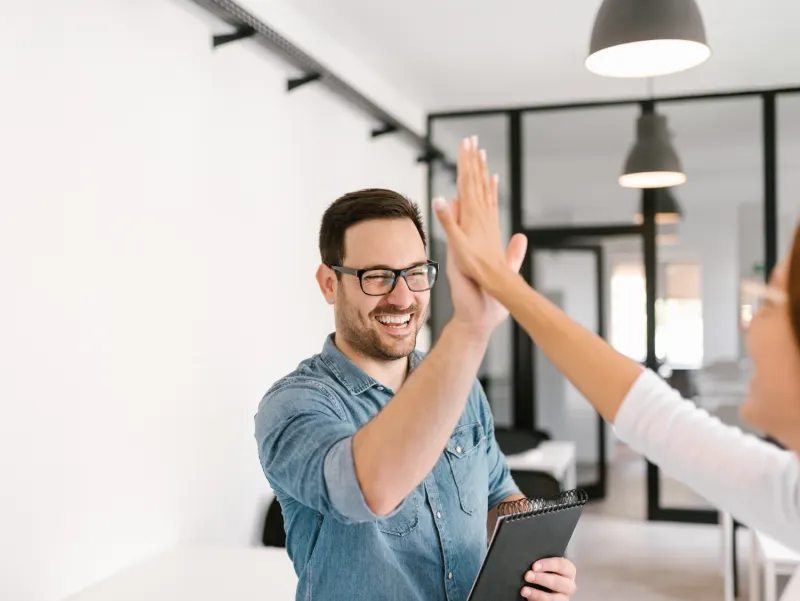 Man in glasses and a denim shirt happily gives a coworker a high five while holding a notepad.