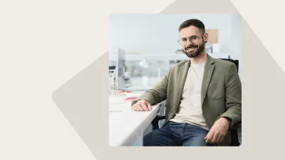 Man with glasses and a beard wearing a green jacket and jeans, smiling while sitting at a desk in an office.