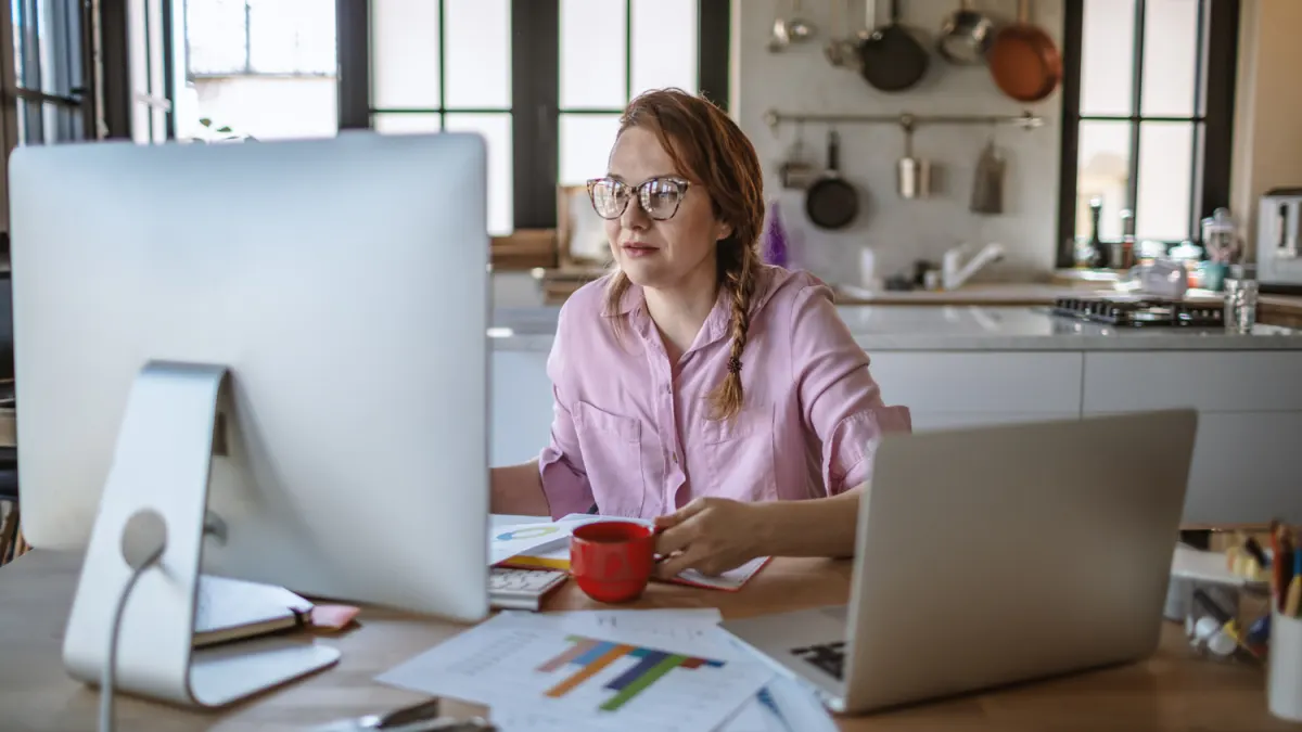 Woman with glasses and braided hair working at a desk with a desktop computer, laptop, and red mug.