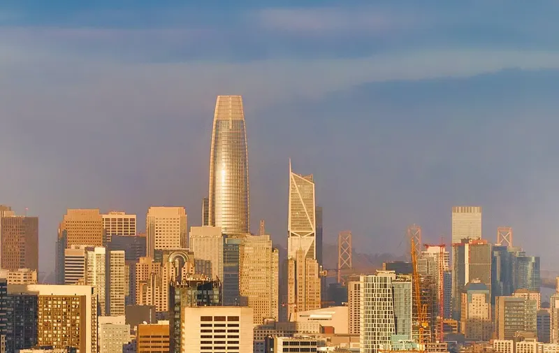 The San Francisco skyline at sunset, featuring the Salesforce Tower and other skyscrapers, with a bridge in the distance.