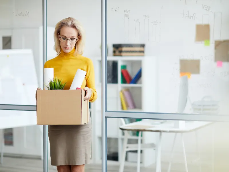 Woman in a yellow sweater holding a box of office supplies, including a plant and rolled papers, looking down.