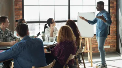 A Black male presenter stands by a whiteboard gesturing toward it with both hands as colleagues sit at a table facing him.