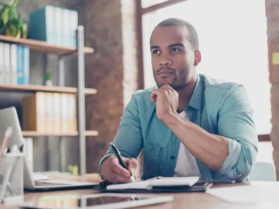 A man in a teal shirt sits at a desk with his hand on his chin, looking thoughtfully into the distance while holding a pen.