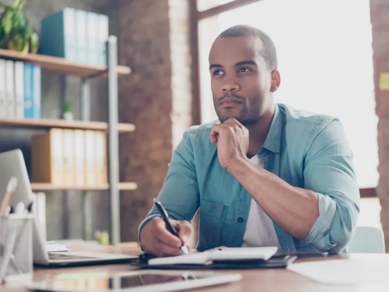 A man in a teal shirt sits at a desk with his hand on his chin, looking thoughtfully into the distance while holding a pen.