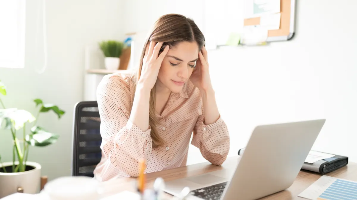 Woman sits at a desk with her eyes closed and hands on her temples, looking stressed while working on a laptop.