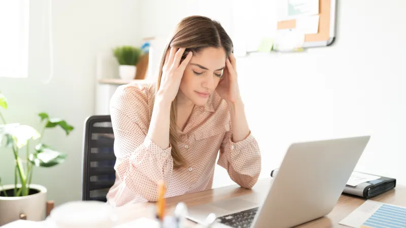 Woman sits at a desk with her eyes closed and hands on her temples, looking stressed while working on a laptop.