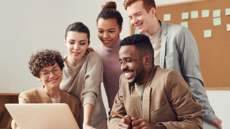 A diverse group of five colleagues collaborate around a laptop, smiling and engaged by the screen.