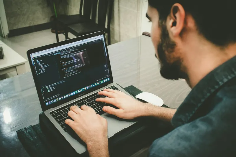 Close-up of a bearded man typing on a MacBook Pro with code on the screen, a white mouse nearby.