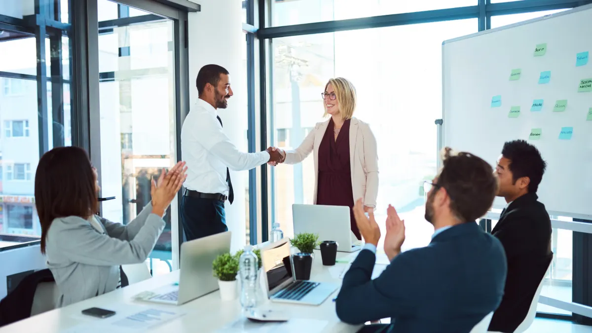 A sales team applauds in a glass-walled meeting room as two colleagues shake hands over a promotion