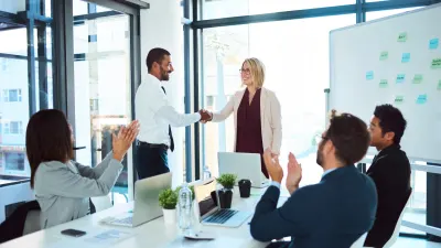 A man and woman shake hands in an office conference room while colleagues clap, a whiteboard with sticky notes behind them.