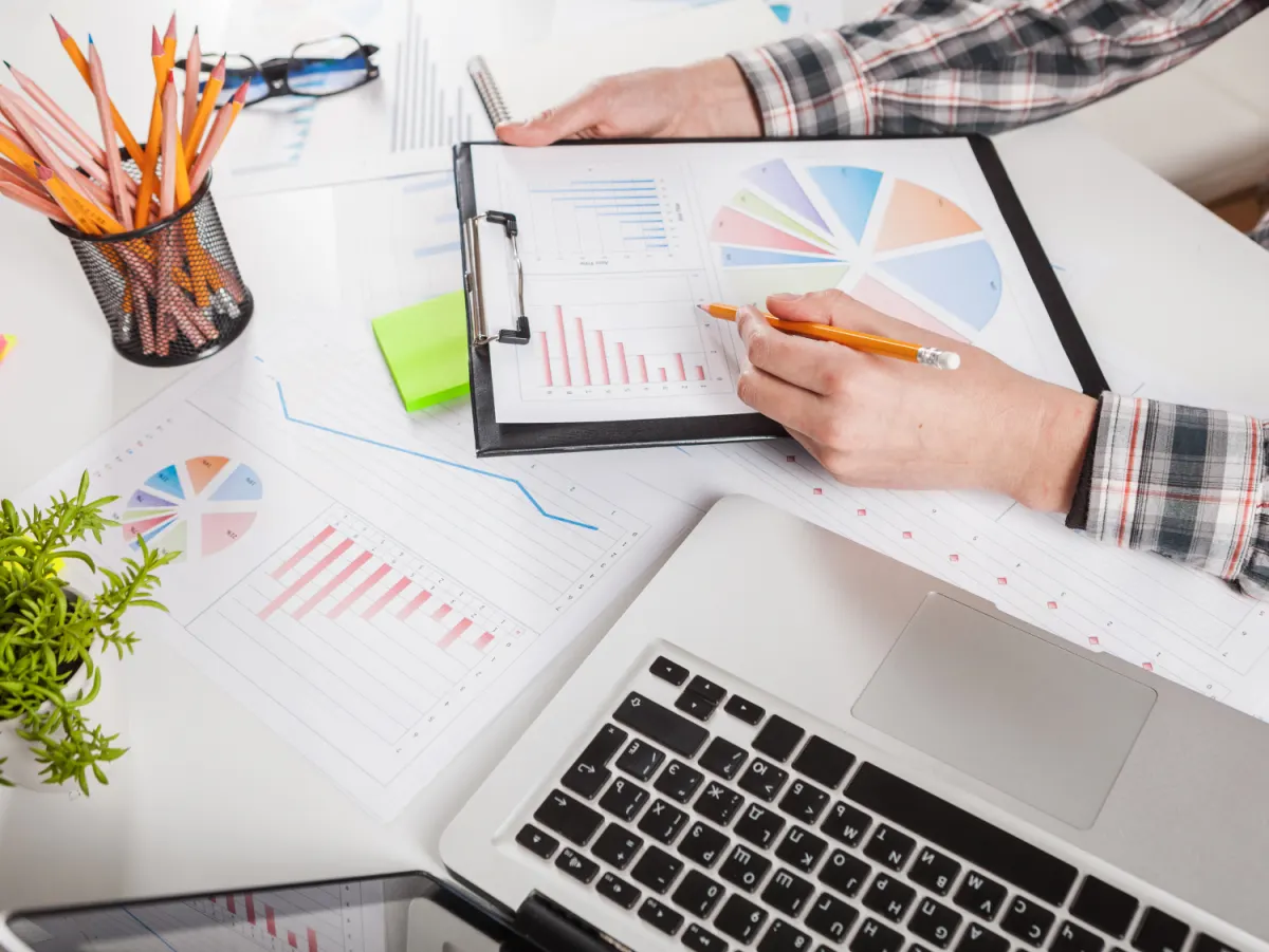 Person in a plaid shirt analyzing charts and graphs with a laptop, pencils, and a plant on the desk.