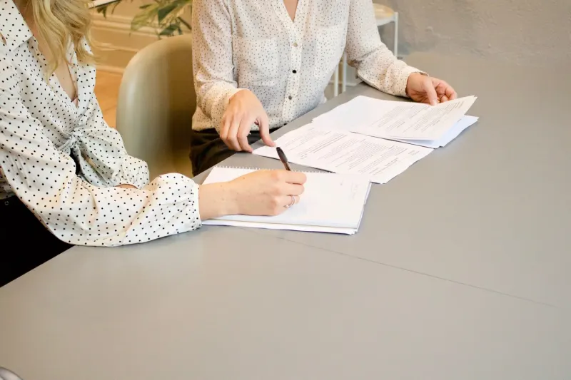 Two people sitting at a table, one writing in a notebook while the other points to documents.