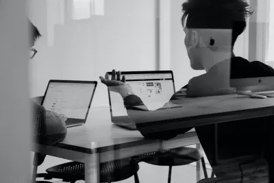 Two people in an office collaborate around laptops, one gesturing to a screen; the image is black and white.